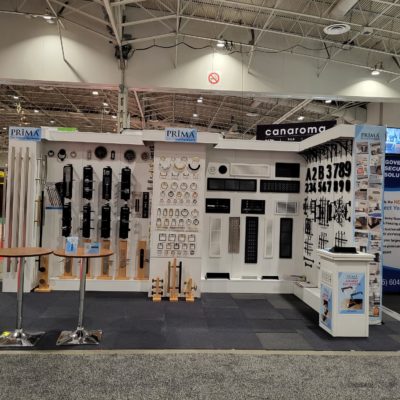 A trade show booth displays various metal hardware, including door handles, knobs, numbers, and modern fixtures arranged on white panels. Signs read PRIMA and canaroma above the booth.