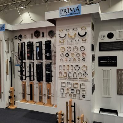 A display wall at a trade show showcases various decorative hardware, including door handles, cabinet pulls, and knobs in different shapes, sizes, and finishes under a PRIMA Decorative Hardware sign.