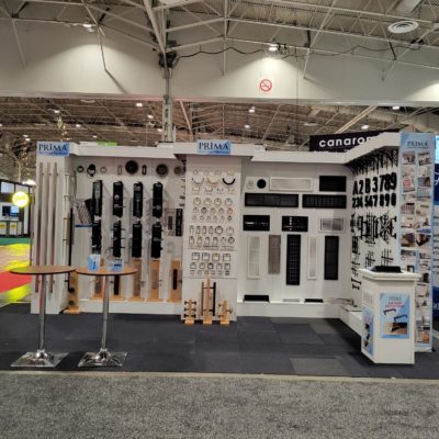 Exhibition booth displaying various hardware products including handles, dials, numbers, and panels on a white wall, with brochures and stools in front. The booth is labeled Prima.