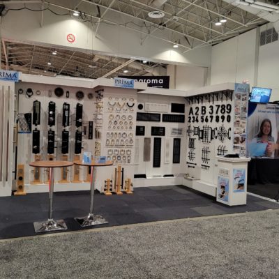 Trade show booth displaying metal hardware, house numbers, door handles, and locks on white walls. A round table with brochures is in front, and a TV screen with a woman holding a blue folder is on the right.