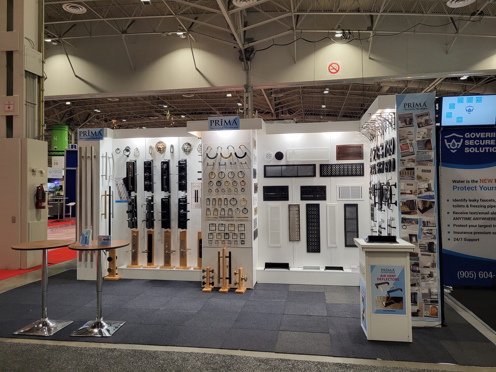 A trade show booth displays various door hardware and security products on white walls, including handles, locks, and panels, with PRIMA branding and informational signs visible.