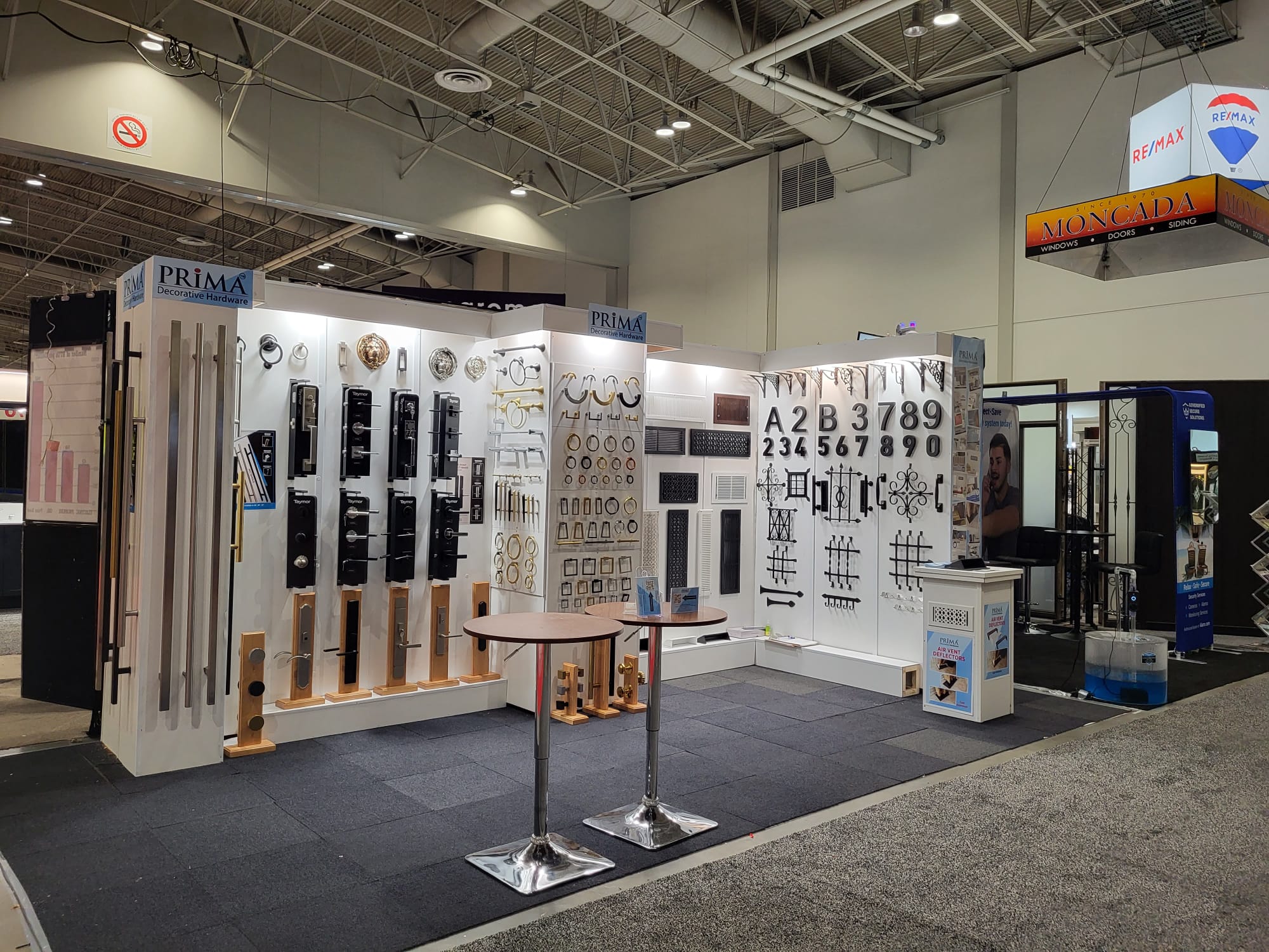 Trade show booth displaying metal hardware, including door handles, railings, house numbers, and decorative elements on white panels. Two round tables with metal bases are in front of the display. Signs read PRIMA.