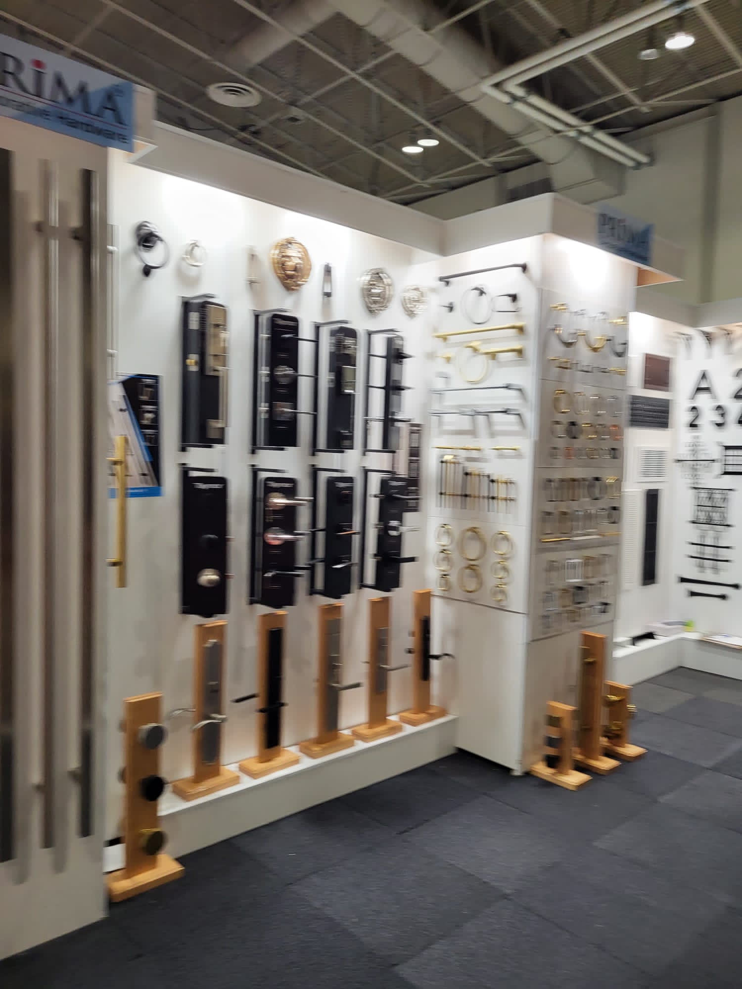 Display booth at a trade show featuring various metal door handles, locks, and hardware mounted on white panels, with some items displayed on wooden stands. The exhibit has a modern, organized appearance.
