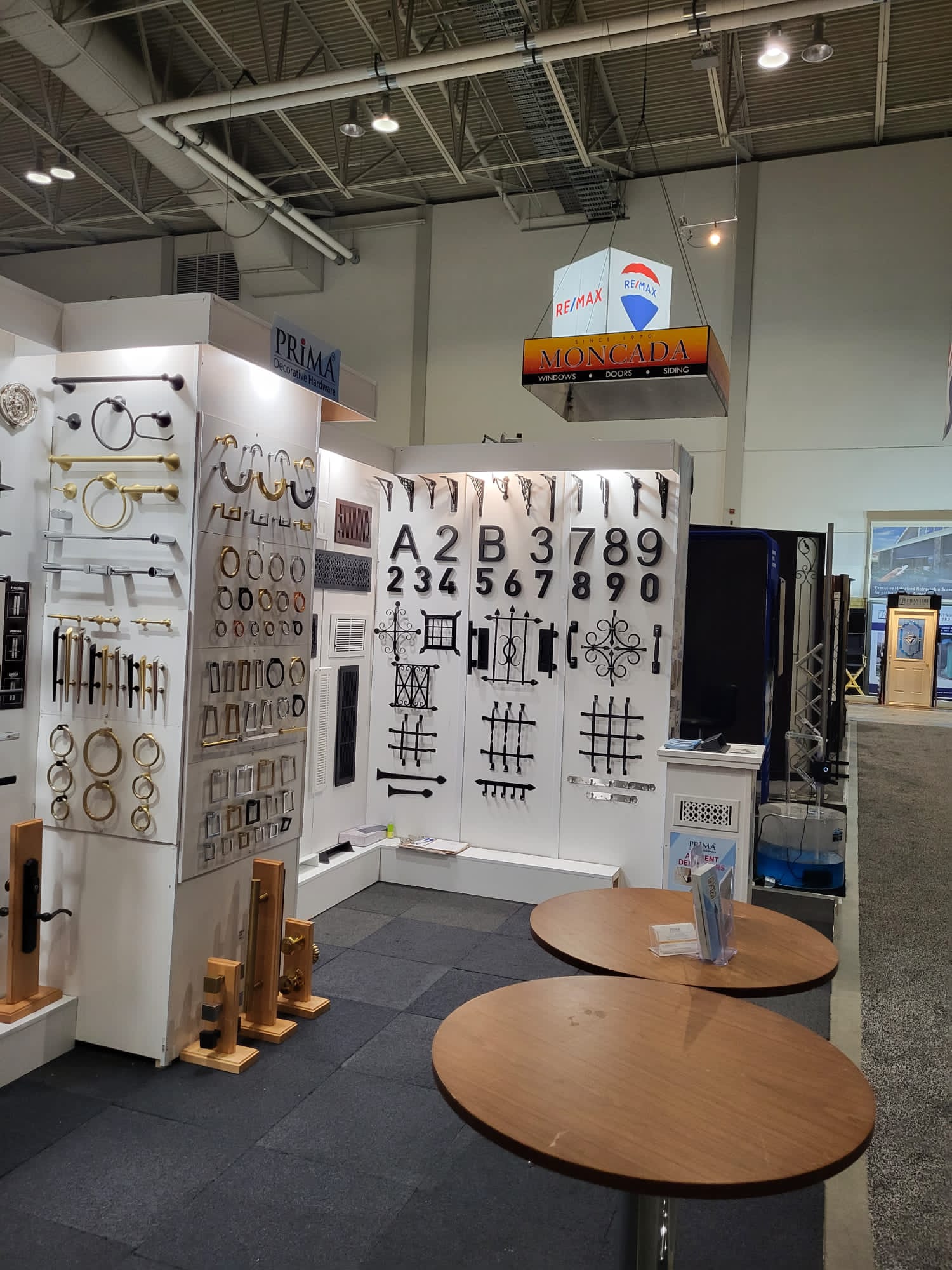 Display booth at a trade show featuring metal hardware, house numbers, handles, and decorative grilles on white panels, with two round wooden tables and business cards in the foreground. Ceiling lights and signs are visible above.