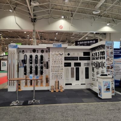 A trade show booth displays various cabinet hardware, including handles, knobs, and numbers, neatly arranged on white panels, with two small round tables and a brochure stand in front.