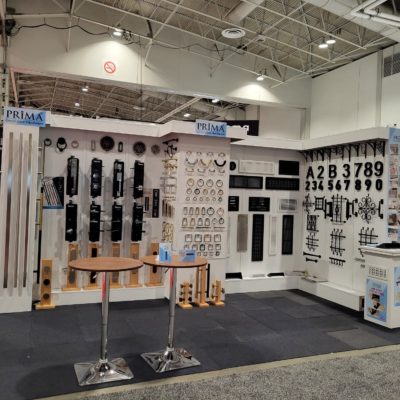 Trade show booth displaying various door hardware, handles, house numbers, and metal fixtures on white walls. Two round tables with chrome bases are in front of the display. The booth is well-lit and organized.