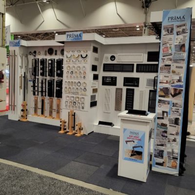 Trade show booth displaying a variety of hardware products on white walls, including vents, grilles, and knobs. The booth has “Prima” branding and informational banners, with a small counter at the front.