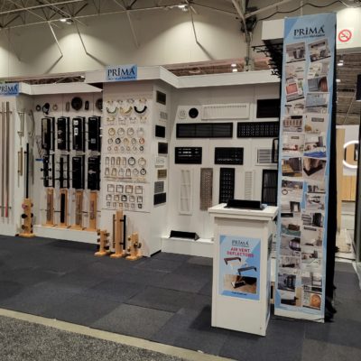 A trade show booth displays various HVAC vents, grilles, and registers mounted on white walls, with product samples on stands and promotional posters branded “PRIMA VENT” throughout the exhibit.