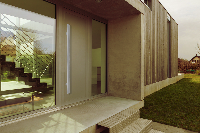 Modern house entrance with large glass door and window, visible metal staircase inside, wooden panel exterior, concrete steps, and green lawn on the side. The scene is quiet and contemporary.