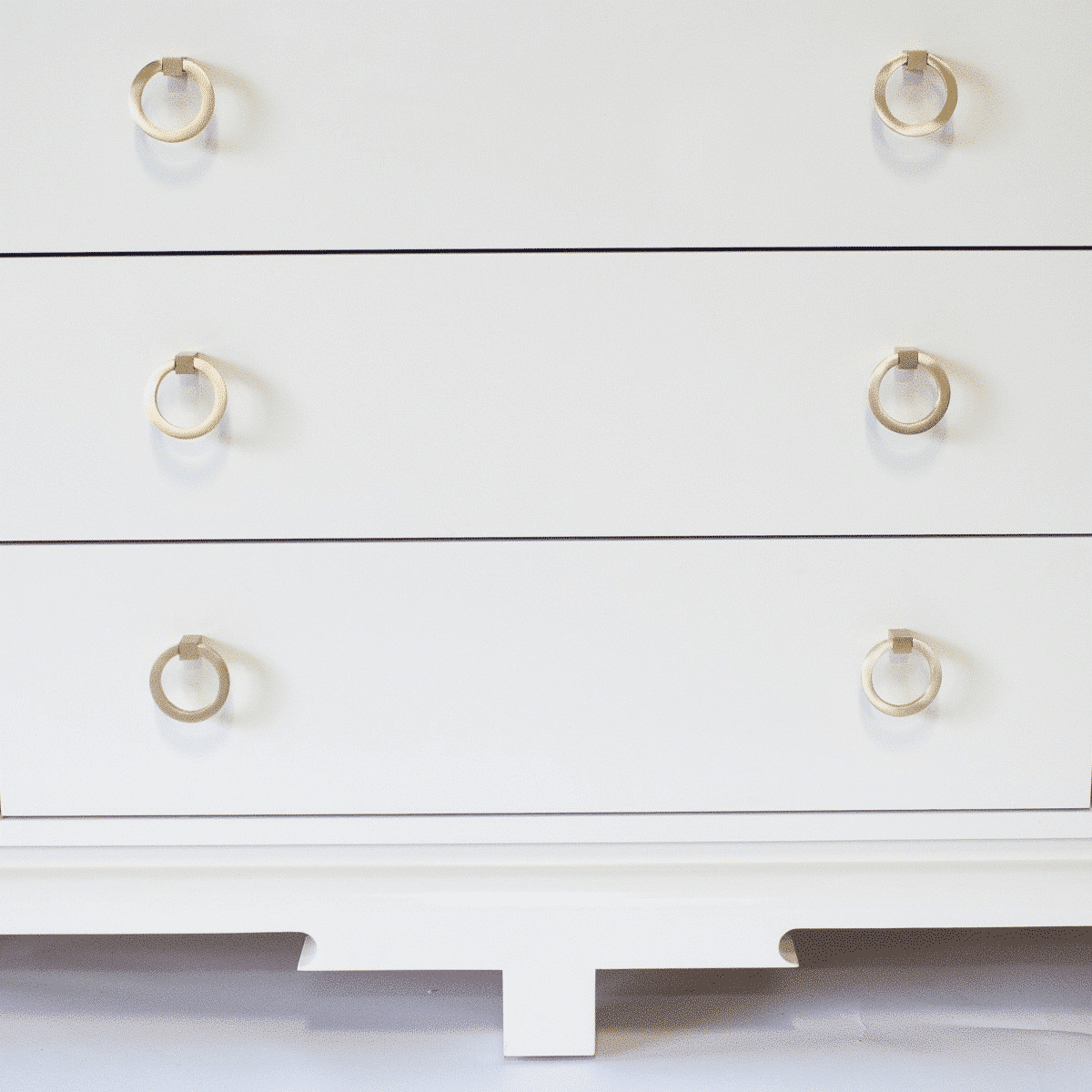 A close-up of a white three-drawer dresser featuring Prima Decorative Hardware's Ring Pulls Round 2″ in brushed brass, set against a plain background. The dresser showcases a simple, modern design with clean lines.