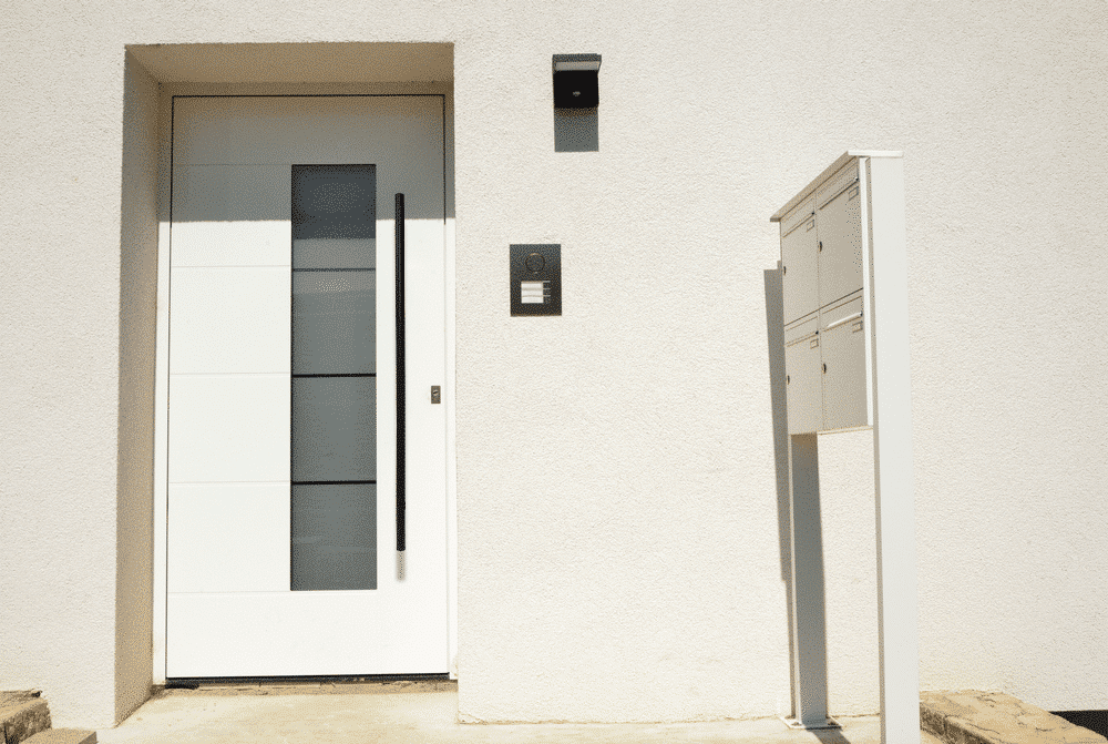 A Prima Decorative Hardware white front door features a vertical stainless steel handle, a compact two-compartment mailbox post, and an intercom plus light fixture on a minimalist white exterior wall.