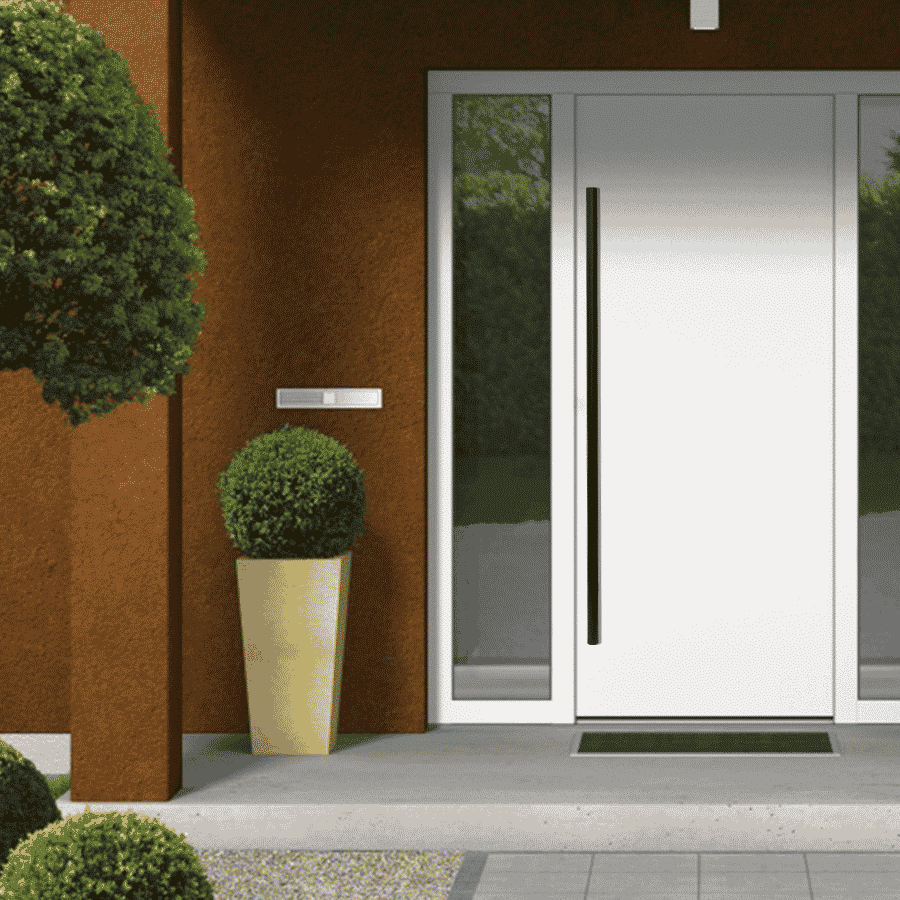 A Prima Decorative Hardware modern white front door with a tall stainless steel handle, mounted in a brown exterior wall. Next to it is a large potted plant, a mailbox above the plant, and a green doormat in front.