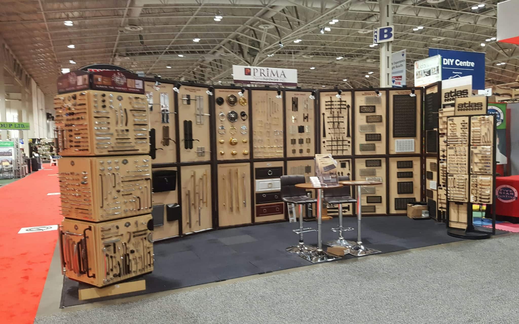Trade show booth displaying a large variety of cabinet handles, knobs, and hardware mounted on wooden boards, with two tall chairs and a small table in front. The area has bright lighting and other booths in the background.