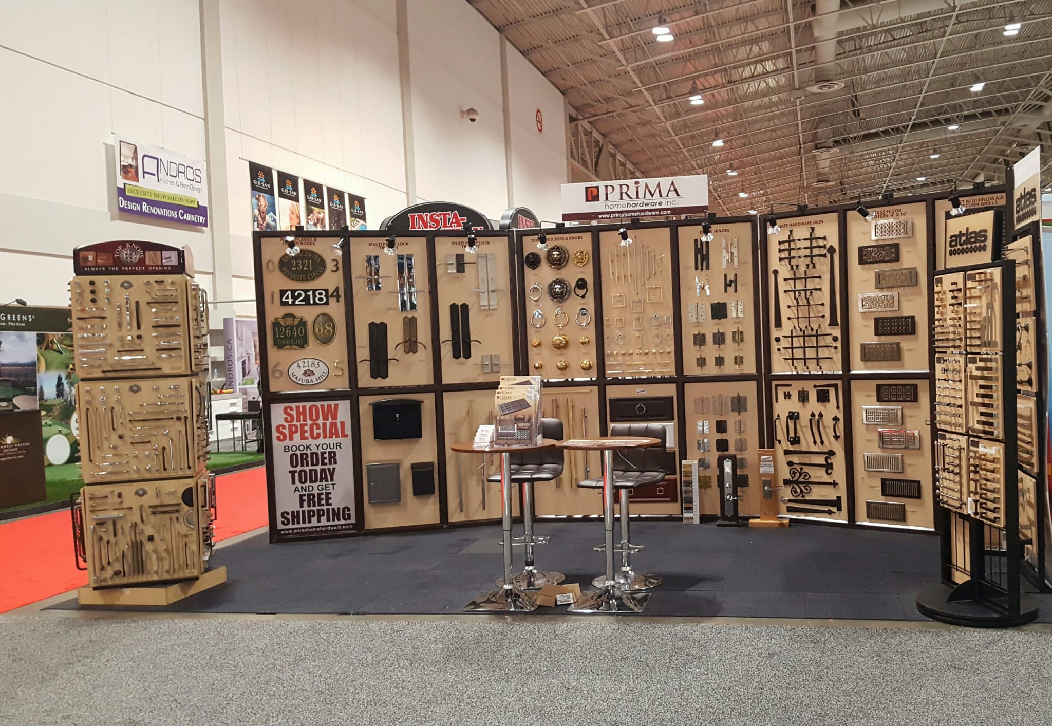 A trade show booth displays various cabinet handles, knobs, and hardware on panels and stands. Two tall chairs and a small table are in the center. Signs advertise free shipping and special show offers.