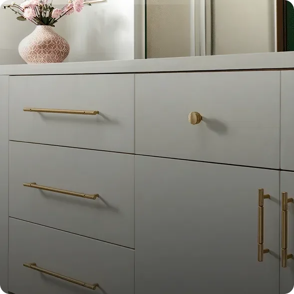 A white cabinet with gold handles and knobs, featuring drawers and doors. A pink vase with flowers sits on the countertop above the cabinet.
