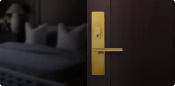 A close-up of a gold door handle and lock on a dark wooden door, with a softly blurred bedroom featuring a bed and cushions visible in the background.