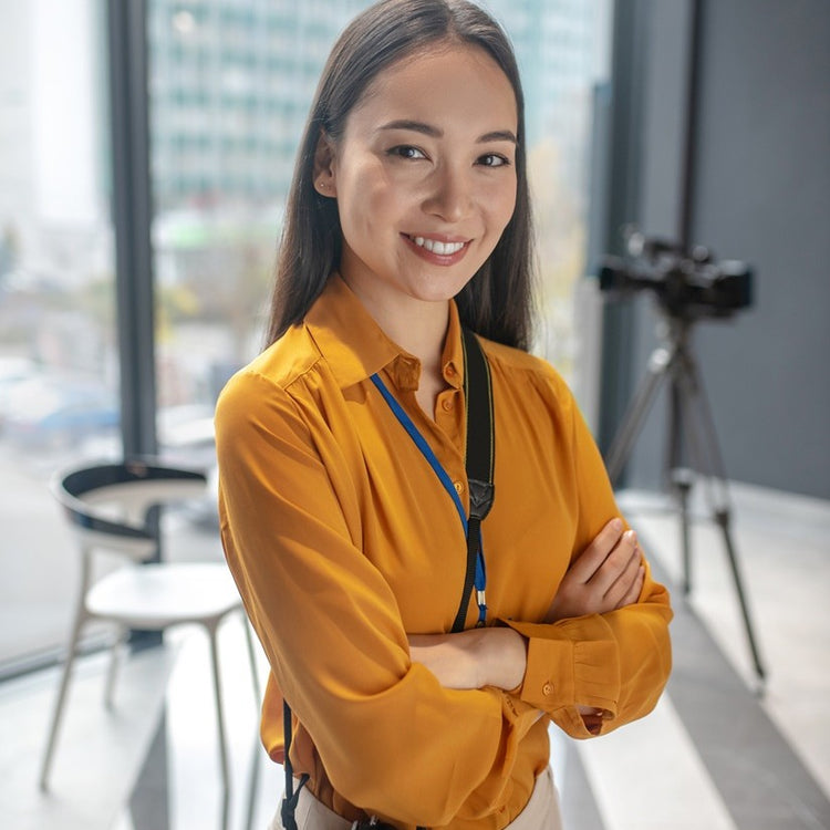 A woman with long dark hair, wearing a mustard yellow blouse and a lanyard, stands smiling with arms crossed in a modern, well-lit office. A camera on a tripod is visible in the background.