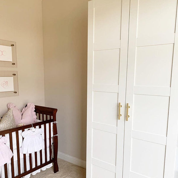 A cozy nursery with a dark wooden crib decorated with pink pillows and baby clothes, next to a large white wardrobe with gold handles against a beige wall.