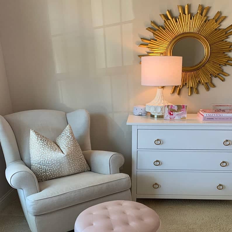 A cozy corner with a cream armchair and ottoman, a white dresser featuring Prima Decorative Hardware's Ring Pulls Round 4", a lit lamp, books, and a sunburst mirror on the wall—all softly illuminated.
