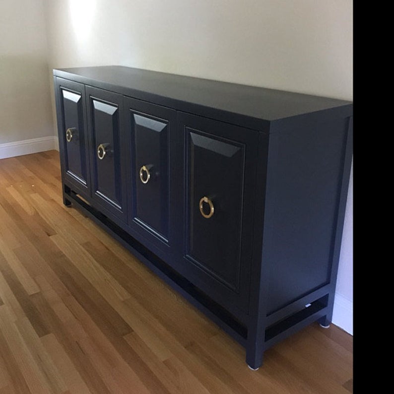 A sleek black sideboard with four glossy cabinet doors, each featuring a Prima Decorative Hardware Ring Pulls Round 4" handle, stands against a light wall on a wooden floor.