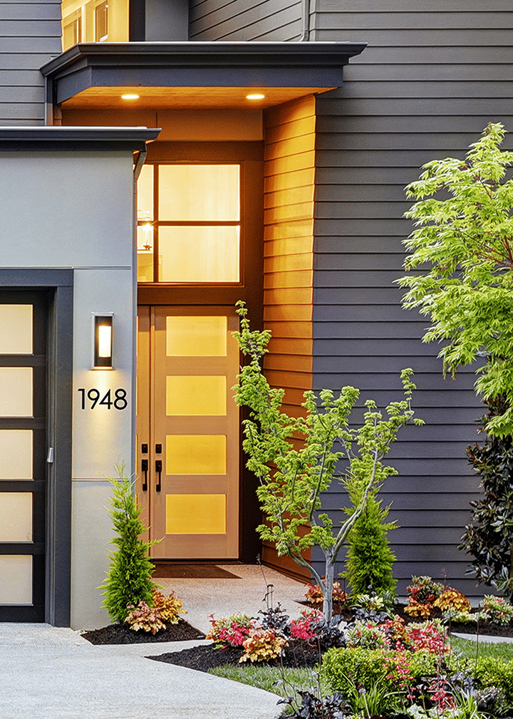 Modern house entrance with frosted glass front door, large windows, and black siding. The house number 1948 is displayed beside the door. Landscaping includes neatly trimmed shrubs, trees, and colorful flowers.