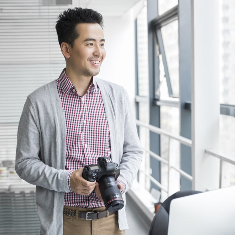 A smiling man in a gray cardigan and checkered shirt holds a professional camera while standing in a bright, modern office with large windows.