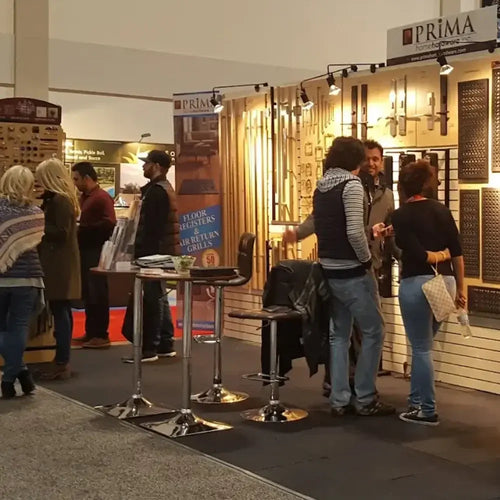 A trade show booth displays decorative floor registers and grilles on illuminated wall panels. Several people are standing, talking, and browsing products. A sign above the booth reads PRIMA.