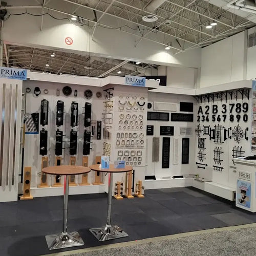 A trade show booth displays various metal hardware, including door handles, numbers, vents, and decorative grilles, organized neatly on white walls. Two round tables with chrome bases stand in the foreground.