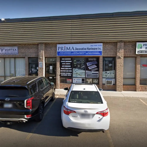Two cars parked in front of a brick strip mall storefront with a sign reading “PRIMA Decorative Hardware Inc.” The entrance has large windows displaying product images and business hours.