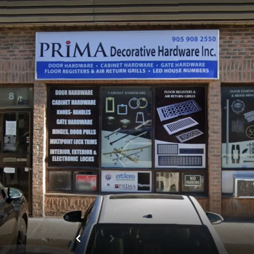 Storefront of PRIMA Decorative Hardware Inc. with signs advertising door and cabinet hardware, locks, handles, registers, and house numbers. Glass windows display images of hardware products and company information.