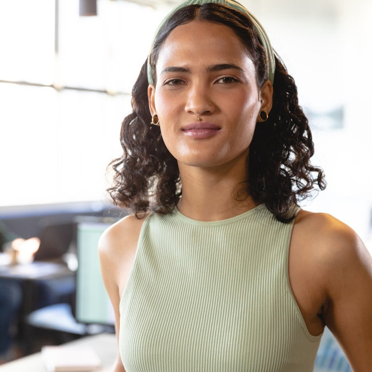 A person with curly dark hair, wearing a light green sleeveless top and headband, stands indoors with a soft smile, in a brightly lit room with blurred background.