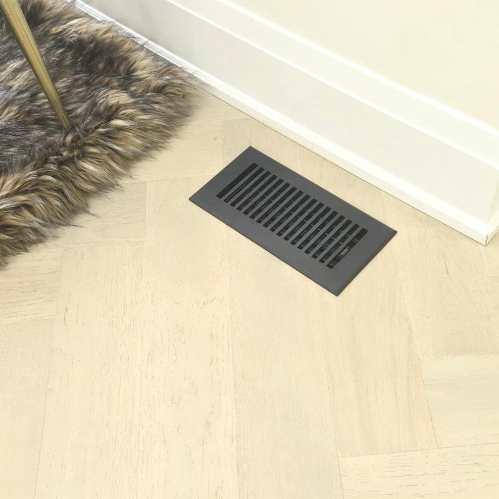 Black floor vent on a wooden floor with a fur rug partially covering it.